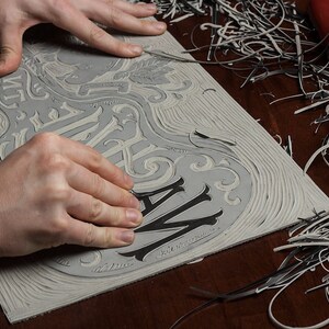 May include: A close-up of a person's hands carving a wooden block with a design that says "The Way" in a decorative font. The block is surrounded by wood shavings.