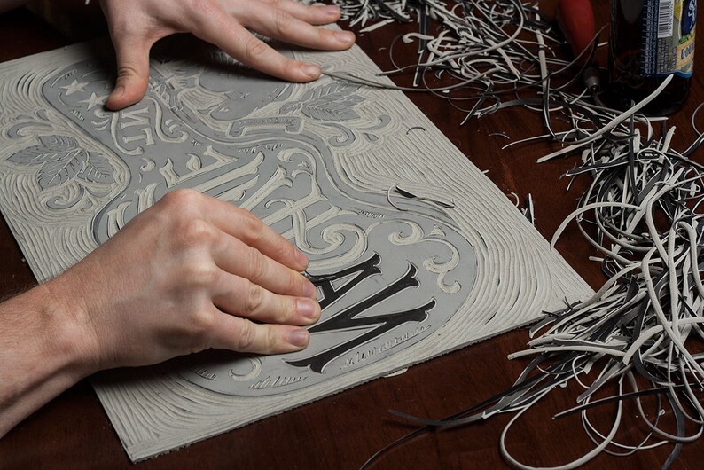 May include: A close-up of a person's hands carving a wooden block with a design that says "The Way" in a decorative font. The block is surrounded by wood shavings.