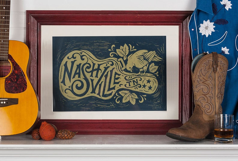 May include: A framed print with a gold design of a guitar shape with the words "Nashville TN" and a bird inside the guitar shape. The print is on a dark blue background and is framed in a dark red wood frame.