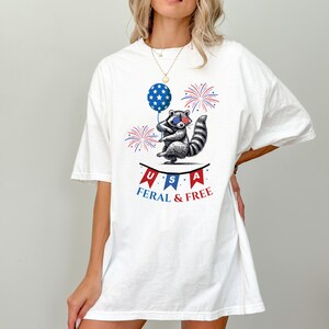 May include: White t-shirt with a graphic of a raccoon wearing sunglasses and holding a red, white, and blue balloon. The raccoon is standing on a banner that says "USA FERAL & FREE".