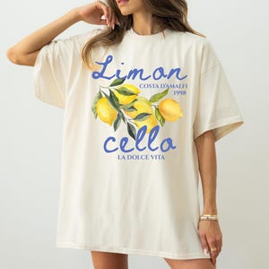 May include: A white oversized t-shirt with a graphic print of lemons and the text "Limon cello Costa d'Amalfi 1998 La Dolce Vita" in blue.