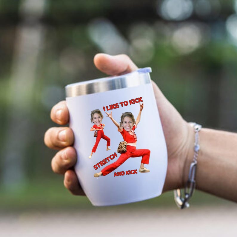 May include: White insulated tumbler with a silver rim and lid. The tumbler features a graphic of a person in a red outfit with the text "I LIKE TO KICK, STRETCH, AND KICK". The tumbler is held in a hand.