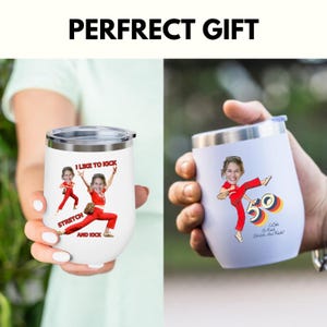 May include: Two white insulated wine tumblers with silver lids. One tumbler has a cartoon image of a person in a red outfit with the text "I LIKE TO KICK, STRETCH AND KICK". The other tumbler has a similar image with the number "50" and the text "I Like To Kick, Stretch and Kick!"