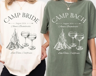 May include: Two white t-shirts with a campfire and glasses design. The text on the shirts reads "Camp Bride" and "Camp Bach" with the date "August 2024" and "Anna's Bachelorette" underneath. The text "Lake Tahoe, California" is at the bottom of the shirts.