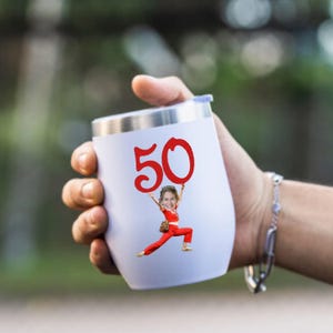 May include: A white insulated tumbler with a silver rim and a blue lid. The number "50" in red is printed on the tumbler, with a cartoon image of a person holding the number. The tumbler is held in a hand.