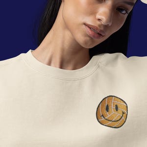 May include: A cream colored sweatshirt with a yellow smiley face embroidered on the left side of the chest. The smiley face is made of a volleyball.