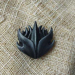 May include: A dark gray, stylized floral-shaped object with pointed petals. The object has a textured surface and is set against a neutral, woven background. The design is reminiscent of a stylized flower or flame.