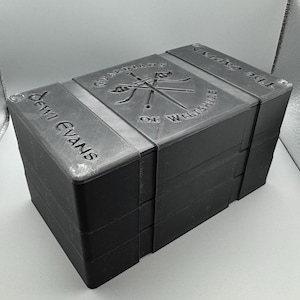 Könnte beinhalten: Eine schwarze, rechteckige Box mit dem Text "Dewi Evans" an der Seite. Die Oberseite der Box zeigt die Worte "Guardians of Wiltshire" und "The Captain" mit einem Emblem aus gekreuzten Schwertern und Hockeyschlägern.