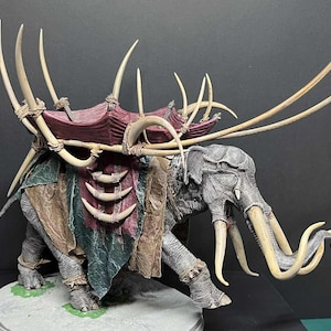 Puede incluir: Modelo detallado de un elefante de guerra, con cuerpo gris, grandes colmillos y una silla de montar burdeos y marrón. El modelo incluye grandes colmillos curvos y elementos decorativos.