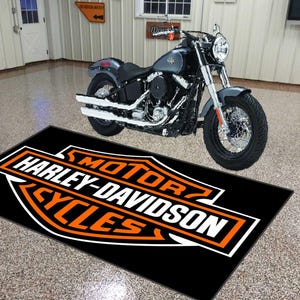 May include: A black Harley-Davidson Cycles floor mat with an orange and white logo is in front of a gray motorcycle. The motorcycle is parked in a garage with a white door and walls.