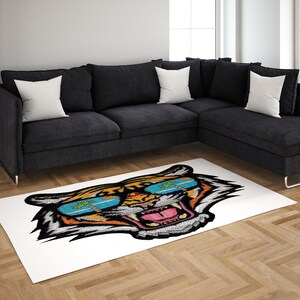 May include: A rectangular white rug featuring a colorful tiger head design wearing sunglasses. The rug is placed on a wooden floor in front of a black sectional sofa with white throw pillows. The tiger's sunglasses have a tropical scene.