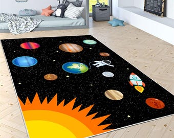 Puede incluir: Una alfombra rectangular negra con un diseño colorido del sistema solar. Planetas, un astronauta y un cohete se representan sobre un fondo estrellado. Un gran sol amarillo y naranja está en la esquina.