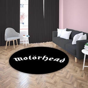 May include: A black, round area rug with the word "motörhead" in white gothic lettering. The rug is placed on a wooden floor in a room with a black sofa, white pillows, and a striped chair.