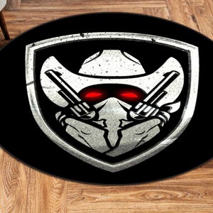 May include: A round black rug featuring a cowboy-themed design. The central graphic is a shield with a cowboy hat, two crossed pistols, and glowing red eyes. The design is in shades of gray and white.