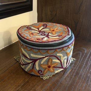 May include: A round, grey fabric storage container featuring a zippered lid and intricate embroidery in shades of orange, cream, and deep red. The floral and vine patterns decorate both the top lid and the vertical sides of the cylindrical case.