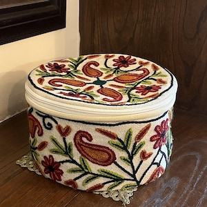 May include: A round, off-white fabric storage container with a zippered lid, featuring colorful embroidered floral and paisley patterns in shades of red, orange, green, and black. The base is accented with a decorative gold-toned trim.