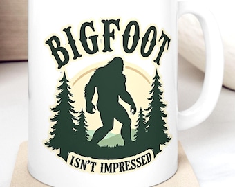 May include: White ceramic mug featuring a Bigfoot silhouette in dark green, with the words "BIGFOOT ISN'T IMPRESSED" in a stylized font. The design includes pine trees and a yellow sun. The mug sits on a beige coaster.