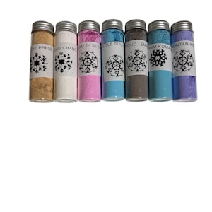 May include: Seven small glass bottles with silver lids, each filled with a different colored powder. The colors include tan, white, pink, light blue, gray, teal, and lavender. Each bottle has a white label with a snowflake design and text.