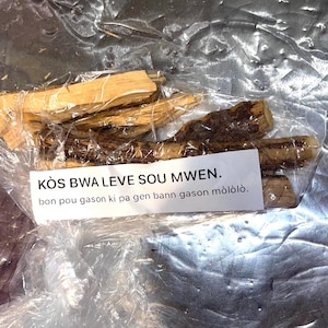May include: A clear plastic bag containing several pieces of brown and light brown wood. The bag has a white label with the text "KÒS BWA LEVE SOU MWEN. bon pou gason ki pa gen bann gason mòlòlò."