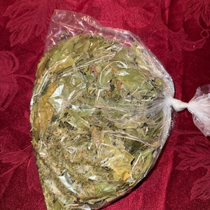 May include: A clear plastic bag filled with dried green leaves. The leaves are various shades of green and brown. The bag is tied at the top with a twist tie. The background is a deep red fabric.