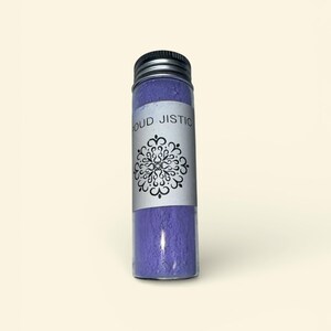May include: A clear glass bottle with a silver lid, filled with a vibrant purple powder. A white label wraps around the bottle, displaying the words "OUD JISTIC" and a black decorative design.