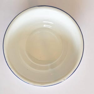 Könnte beinhalten: Eine weiße Emaille-Schüssel mit blauem Rand. Die Schüssel hat ein schlichtes, klassisches Design und eine glänzende Oberfläche. Der Innenraum ist glatt und reflektierend, ideal zum Servieren oder Zubereiten von Speisen.