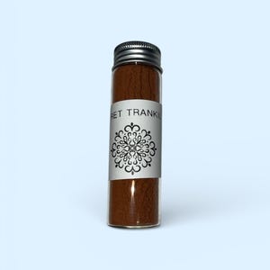 Op de afbeelding: Een klein glazen flesje met een zilveren dop, gevuld met bruin poeder. Een wit etiket met zwarte tekst en een decoratief bloemmotief is om het flesje gewikkeld. De tekst op het etiket luidt "RET TRANKIL".