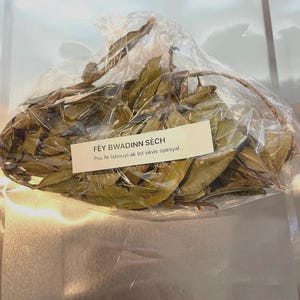 May include: A clear plastic bag filled with dried green leaves, tied with twine. A white label with black text is attached to the bag. The text on the label reads "FÈY BWADINN SÈCH" and "Pou te labouyi ak lot sèvis spesyal."
