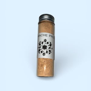 Könnte beinhalten: Ein kleines Glasfläschchen mit silbernem Deckel, gefüllt mit hellbraunem Pulver. Ein weißes Etikett umschließt das Fläschchen und zeigt den Text "MACHE PRE" über einem schwarzen floralen Design. Das Fläschchen steht vor einem hellblauen Hintergrund.