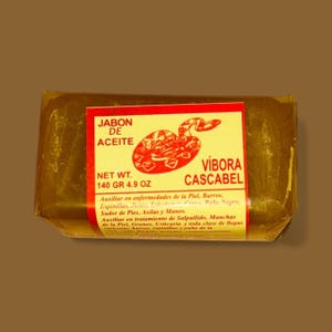 May include: A bar of soap wrapped in clear plastic. The label is red and yellow with the text "JABON DE ACEITE VÍBORA CASCABEL" and a graphic of a snake. The net weight is 4.9 oz (140 grams).