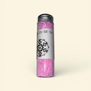 以下が含まれることがあります： ピンク色の粉末が入った、銀色の蓋が付いた円筒形のガラス瓶。瓶には「M DI SE SA」の文字と黒い雪の結晶のデザインが施された白いラベルが巻かれています。瓶は明るいベージュ色の背景に置かれています。