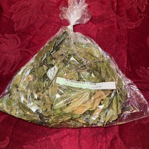 May include: A clear plastic bag filled with dried green leaves, tied at the top. A white label with black text is visible on the bag. The bag is sitting on a red patterned surface.