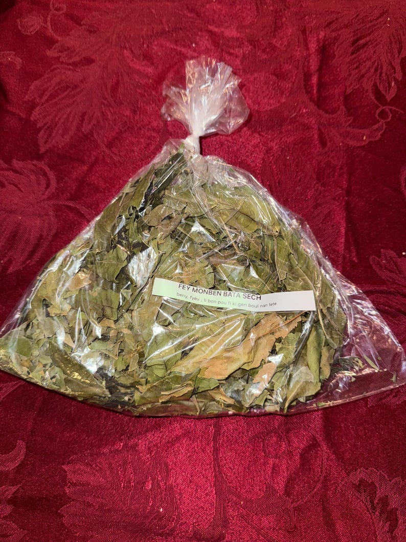 May include: A clear plastic bag filled with dried green leaves, tied at the top. A white label with black text is visible on the bag. The bag is sitting on a red patterned surface.