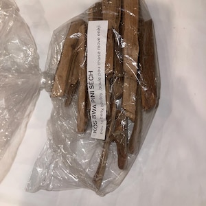 May include: A transparent plastic bag containing dried, brown wooden sticks. A white label with black text is attached to the bag, reading "KOS BWA PINI SÈCH" and other text. The sticks are of different lengths and thicknesses.