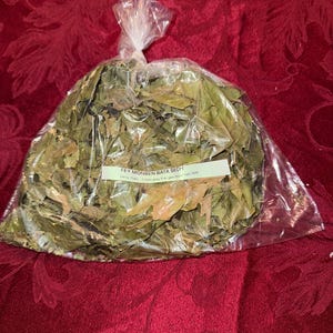 May include: A clear plastic bag filled with dried green leaves. A white label with black text is attached to the bag. The bag is tied at the top. The background is a red, textured fabric.