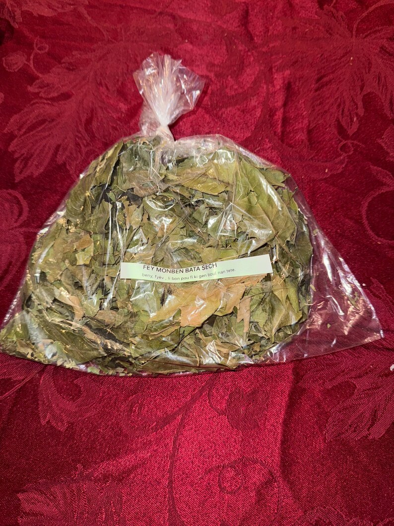 May include: A clear plastic bag filled with dried green leaves. A white label with black text is attached to the bag. The bag is tied at the top. The background is a red, textured fabric.