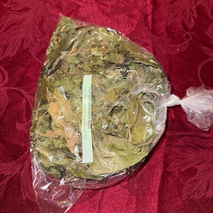 May include: A clear plastic bag filled with dried green leaves, tied at the top. A white label with black text is attached to the bag. The bag is resting on a red patterned surface.