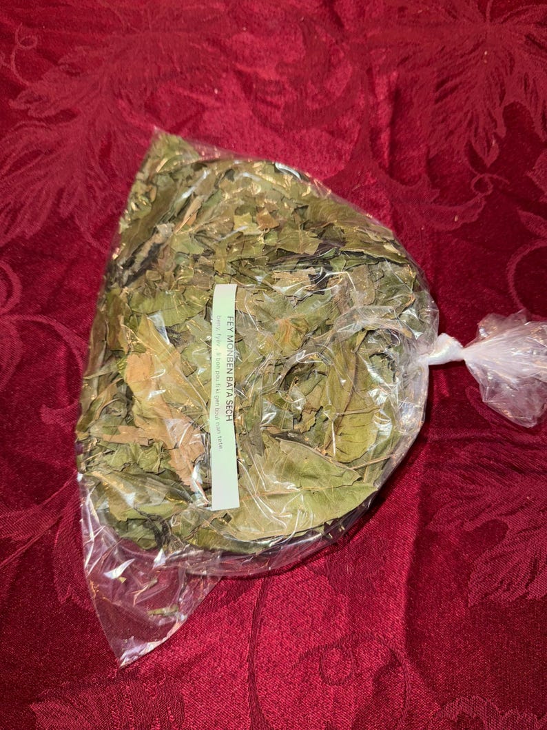 May include: A clear plastic bag filled with dried green leaves, tied at the top. A white label with black text is attached to the bag. The bag is resting on a red patterned surface.