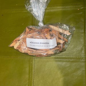 Puede incluir: Una bolsa de plástico transparente llena de pequeñas astillas de madera. La bolsa tiene una etiqueta que dice "KOS BWA BWADOM. bon pou emoyid ak lise nan kol matris."