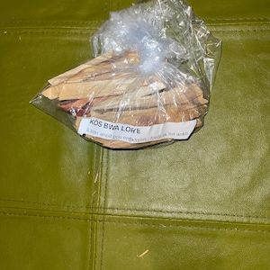 May include: A clear plastic bag filled with thin wooden sticks. The bag has a label that reads "KOS BWA LORE" and "li bon anpil pou enkisyon, fred ak lot anko."