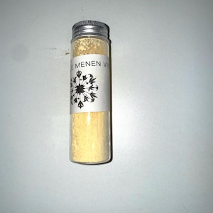 May include: A small, clear glass vial with a silver metal cap, filled with a yellow powder. A white label with black text and a floral design is affixed to the vial. The text on the label reads "E MENEN VI".