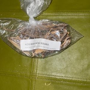 May include: A clear plastic bag filled with dried brown roots. The bag has a white label with the text "KOS BWA KOK SOURIT. bon kont maladi ki ap prodwi nan system."
