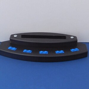 Puede incluir: Un soporte de pl&aacute;stico negro con cinco ladrillos Lego azules en la parte delantera. El soporte tiene una ranura en la parte superior para sujetar un tel&eacute;fono u otro dispositivo.