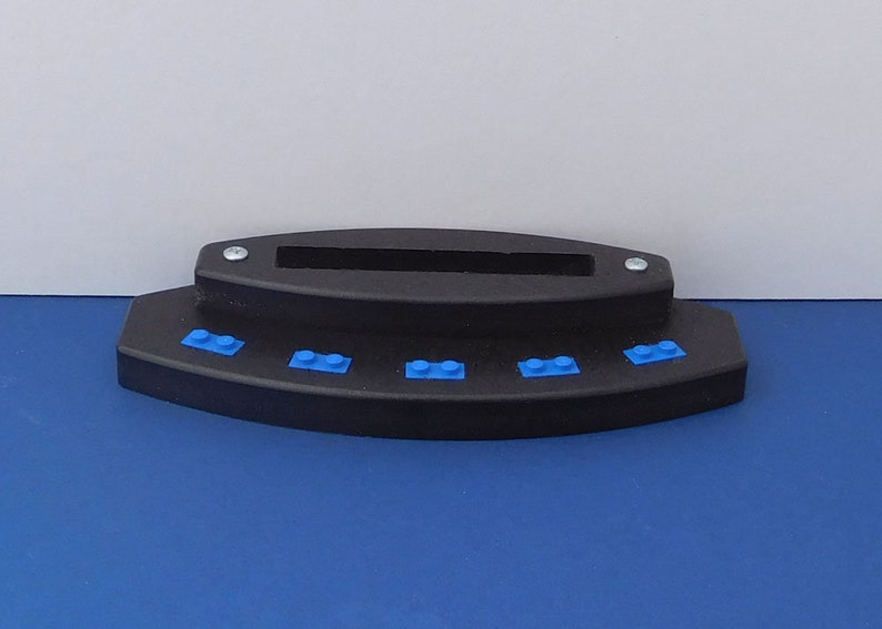 Puede incluir: Un soporte de pl&aacute;stico negro con cinco ladrillos Lego azules en la parte delantera. El soporte tiene una ranura en la parte superior para sujetar un tel&eacute;fono u otro dispositivo.