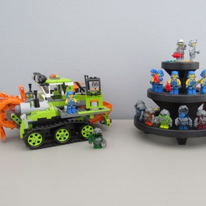 May include: A green and orange toy construction vehicle with a rotating drill and tracks. The vehicle is surrounded by smaller toy figures, including blue and grey figures with various accessories.