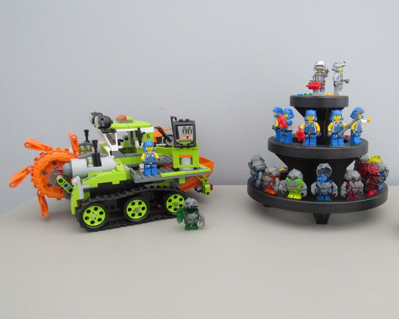 May include: A green and orange toy construction vehicle with a rotating drill and tracks. The vehicle is surrounded by smaller toy figures, including blue and grey figures with various accessories.
