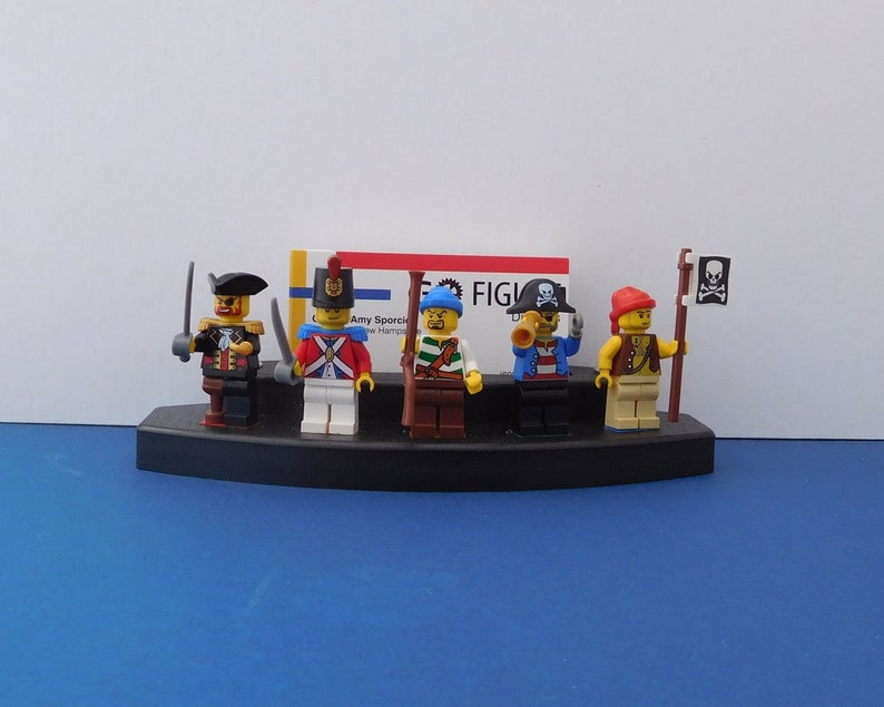 Puede incluir: Cinco figuras Lego en miniatura en un expositor negro. Las figuras son un pirata, un soldado, un pirata con un telescopio, un pirata con una espada y un pirata con una bandera de calavera y huesos cruzados. Las figuras llevan todas ropa y accesorios de diferentes colores.