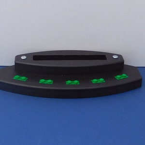Puede incluir: Un soporte de pl&aacute;stico negro con una ranura para sujetar un tel&eacute;fono u otro dispositivo. El soporte tiene una fila de cinco ladrillos Lego verdes en la parte delantera.