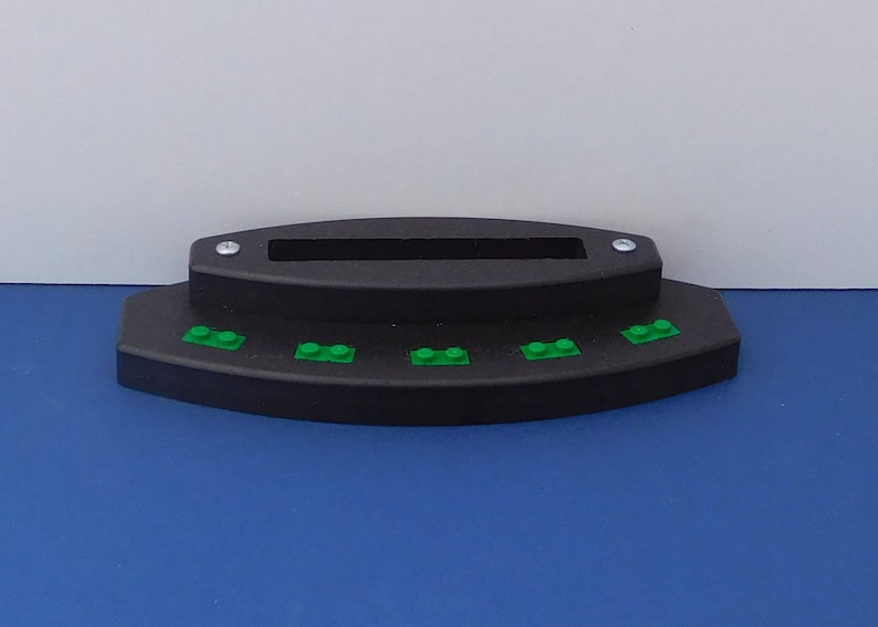 Puede incluir: Un soporte de pl&aacute;stico negro con una ranura para sujetar un tel&eacute;fono u otro dispositivo. El soporte tiene una fila de cinco ladrillos Lego verdes en la parte delantera.