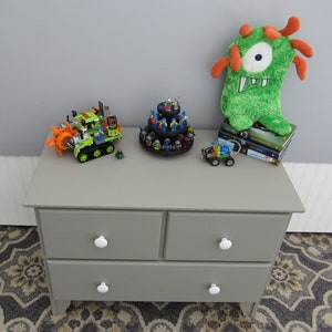 May include: A gray dresser with three drawers and white knobs. On top of the dresser are a green and orange plush monster, a Lego toy vehicle, a Lego minifigure display stand, and a stack of books.